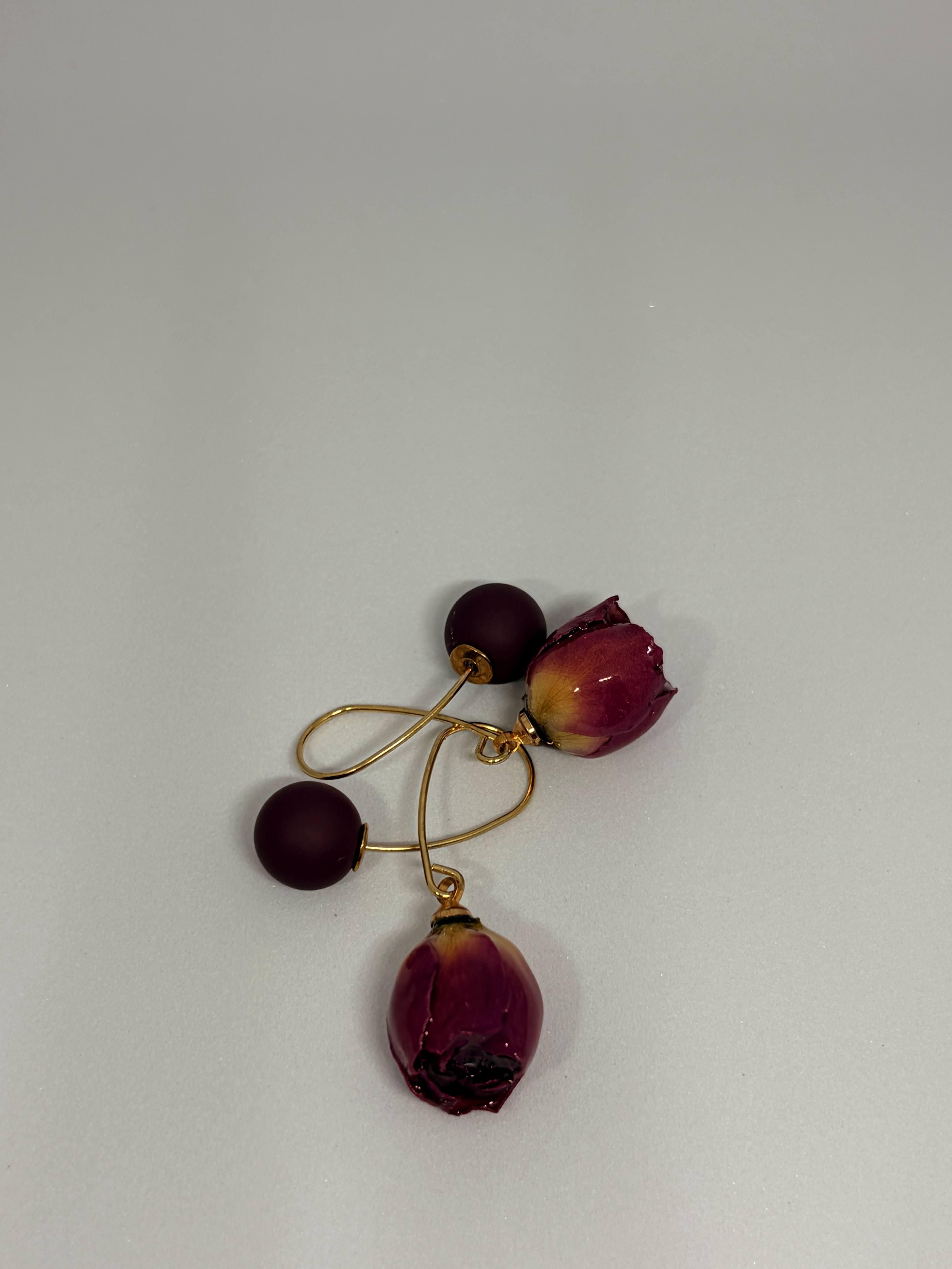 A pair of gold-tone dangle earrings featuring matte dark burgundy beads and dried rosebud charms, displayed on a neutral light gray background.