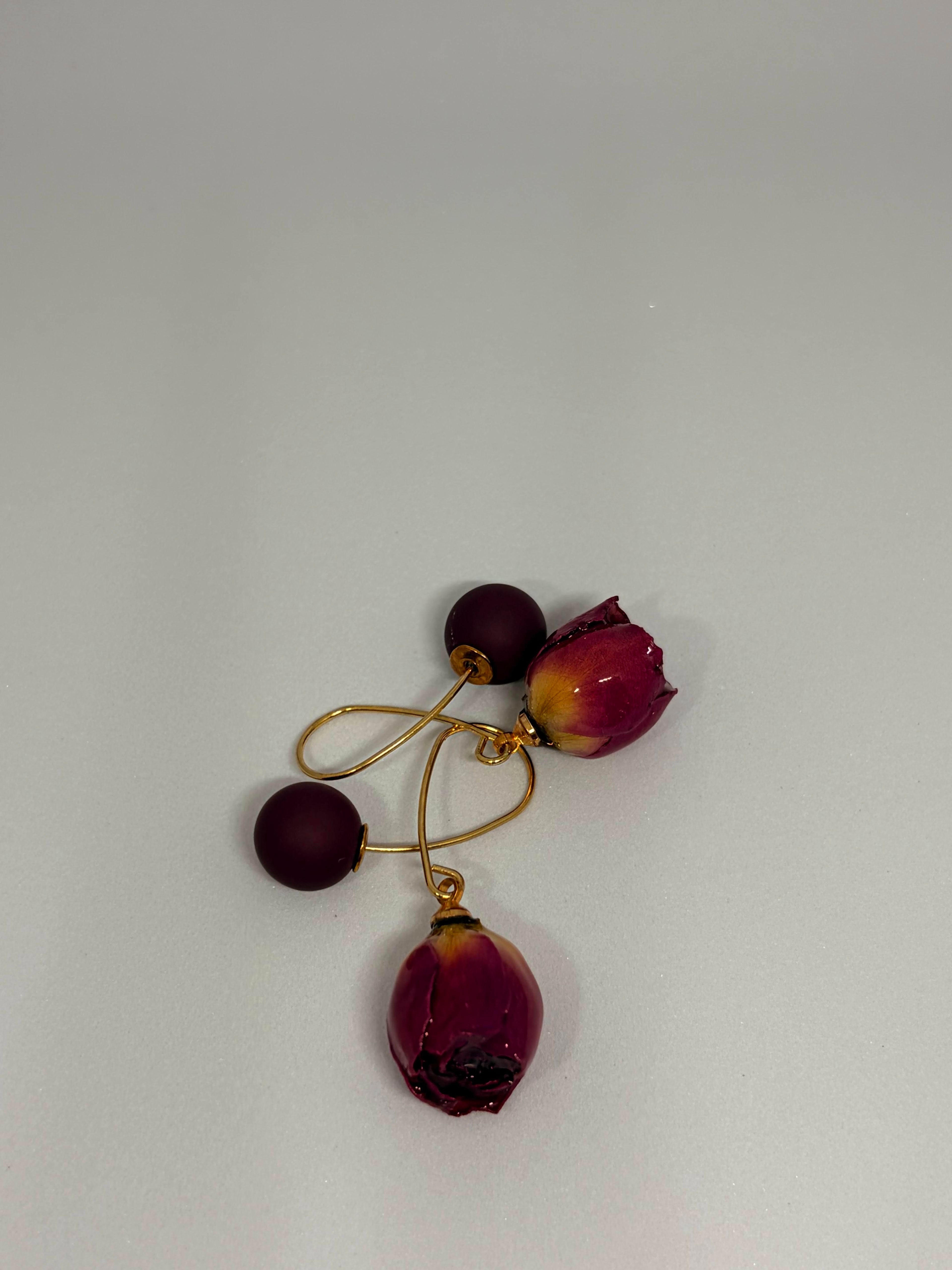 A pair of gold-tone dangle earrings featuring matte dark burgundy beads and dried rosebud charms, displayed on a neutral light gray background.
